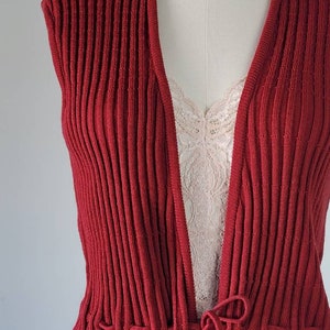 May include: A red ribbed knit sleeveless cardigan with a tie at the waist.