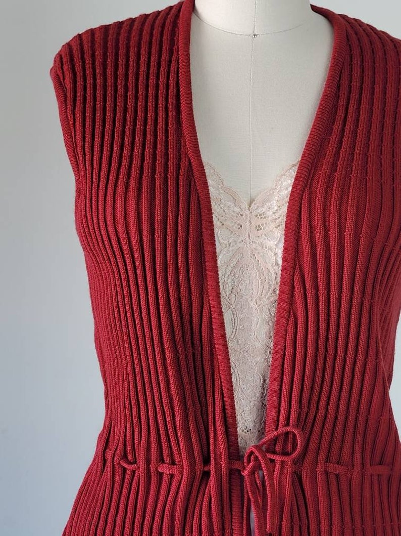 May include: A red ribbed knit sleeveless cardigan with a tie at the waist.