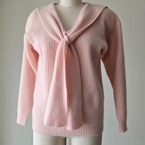 May include: A light pink sweater with a unique scarf-like collar tied in a knot. The sweater has long sleeves and a ribbed texture. The garment is displayed on a mannequin, showcasing its design and fit.