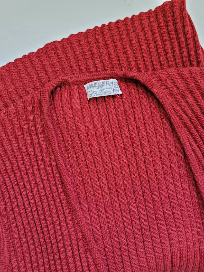 May include: A red ribbed knit cardigan with a V-neck. The label reads "Jaeger" and includes information about the material and care instructions.