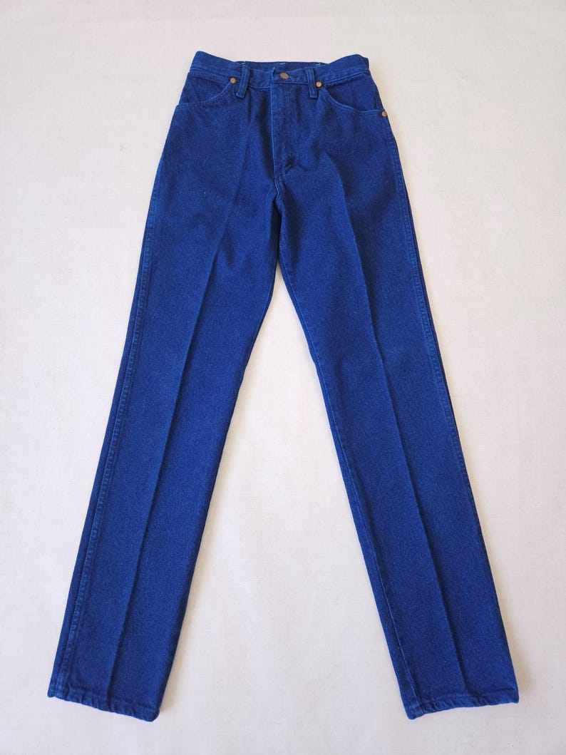 May include: A pair of classic blue denim jeans. The jeans have a straight leg cut, a button and zipper closure, and pockets. The denim is a solid, medium blue color. The jeans are laid flat on a white surface.
