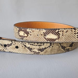 May include: A snakeskin belt with a silver buckle. The belt has a pattern of tan, brown, and black scales. The inside of the belt is light brown. The belt is likely to be made of genuine leather.