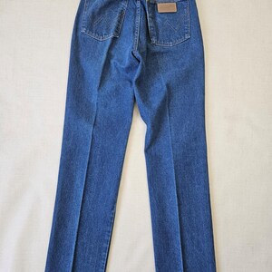 May include: Blue denim jeans with a classic five-pocket design. The jeans feature a brown leather patch on the back pocket and the Wrangler logo. The jeans are laid flat, showcasing the back view.