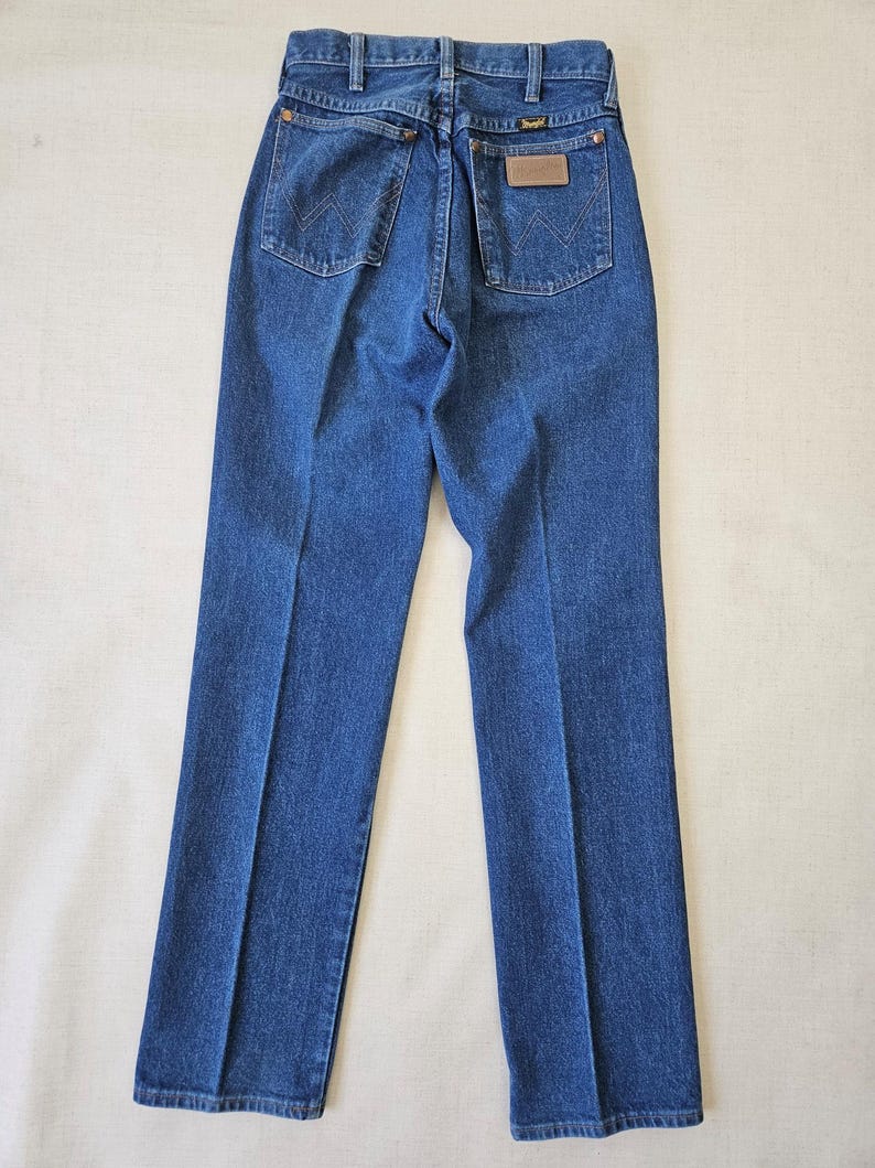 May include: Blue denim jeans with a classic five-pocket design. The jeans feature a brown leather patch on the back pocket and the Wrangler logo. The jeans are laid flat, showcasing the back view.