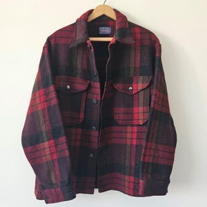 Puede incluir: Una camisa de manga larga con botones y un patrón de cuadros en rojo oscuro, negro y marrón. La camisa incluye un cuello puntiagudo, dos bolsillos en el pecho con solapas abotonadas y una fila central de botones oscuros. Se ve una etiqueta Pendleton.