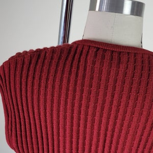 May include: A close-up of a red ribbed knit sweater. The sweater has a round neckline and a smooth texture.