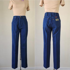May include: A pair of dark blue denim jeans with a straight leg cut. The jeans have a classic five-pocket design, a button and zipper closure, and a brown leather patch on the back pocket. The jeans are displayed on a mannequin.