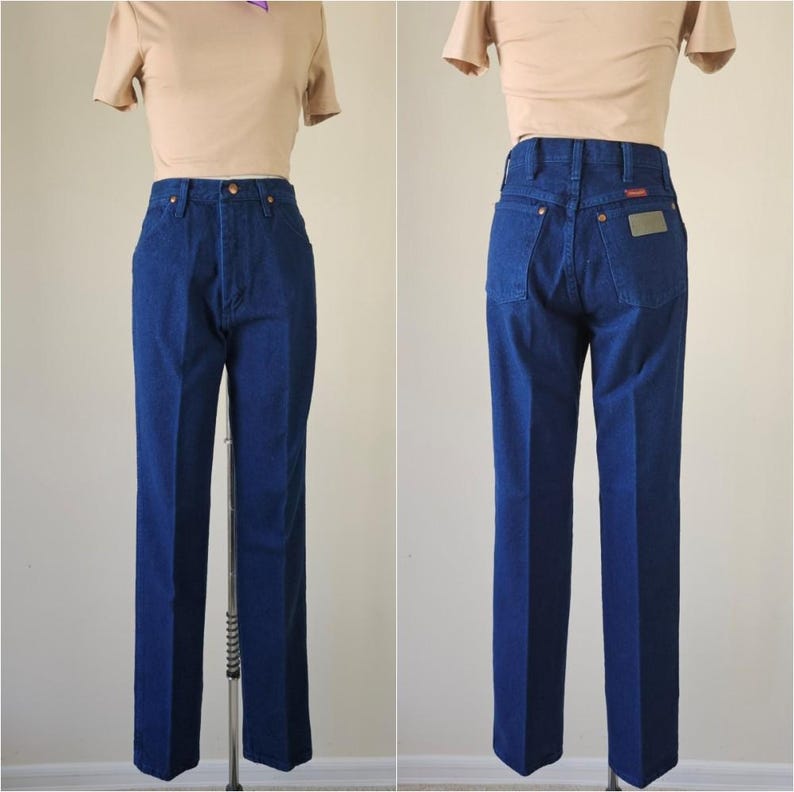May include: A pair of dark blue denim jeans with a straight leg cut. The jeans have a classic five-pocket design, a button and zipper closure, and a brown leather patch on the back pocket. The jeans are displayed on a mannequin.
