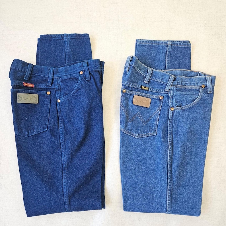 May include: Two pairs of blue denim jeans. One pair is a darker shade of blue, and the other is a lighter shade. Both pairs have the Wrangler logo on the back pocket and a leather patch.