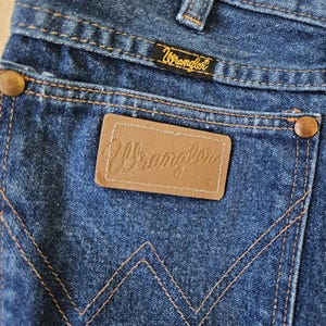 May include: Close-up of blue denim jeans with the Wrangler logo on the back pocket and a small tag. The jeans have copper-colored rivets and stitching. The Wrangler logo is also on a small black tag.