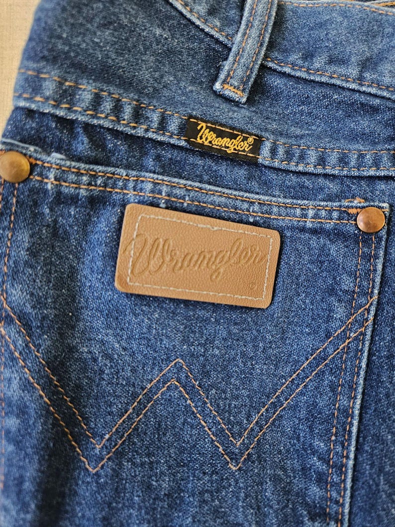 May include: Close-up of blue denim jeans with the Wrangler logo on the back pocket and a small tag. The jeans have copper-colored rivets and stitching. The Wrangler logo is also on a small black tag.