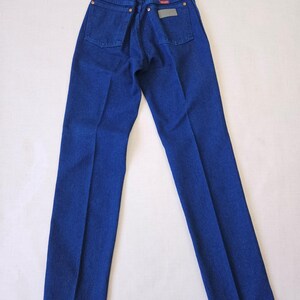 May include: A pair of blue denim jeans, viewed from the back. The jeans have two back pockets, a small rectangular patch, and copper-colored rivets. The denim is a solid, deep blue color.