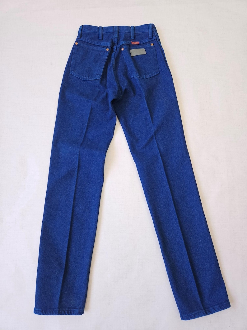 May include: A pair of blue denim jeans, viewed from the back. The jeans have two back pockets, a small rectangular patch, and copper-colored rivets. The denim is a solid, deep blue color.