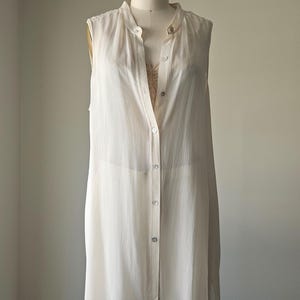 May include: A sleeveless, off-white button-down tunic shirt and matching pants are displayed on a mannequin. The tunic has a mandarin collar and a button-down front. The pants are wide-legged and the fabric appears to be lightweight.