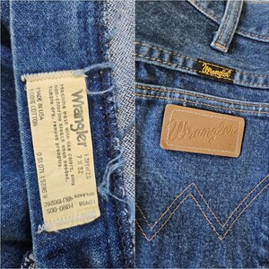 May include: Close-up of blue denim Wrangler jeans. The jeans feature a leather patch with the Wrangler logo, a small tag with the Wrangler brand name, and a care label. The jeans are made of 100% cotton.