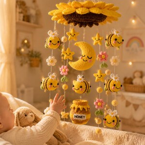 Op de afbeelding: Een gehaakte babykamer mobiel met een grote gele zonnebloem aan de bovenkant, met hangende elementen zoals lachende bijtjes, gele sterren, roze bloemen, een gele halve maan en een honingpot met het woord HONEY op de voorzijde gedrukt.