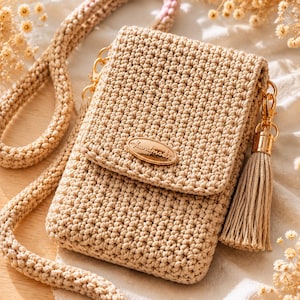 Può includere: Una borsa a tracolla in uncinetto color beige con patta rettangolare e un'etichetta metallica ovale color oro con la scritta handmade. La borsa include una tracolla in uncinetto abbinata e una nappa decorativa beige fissata con elementi dorati.