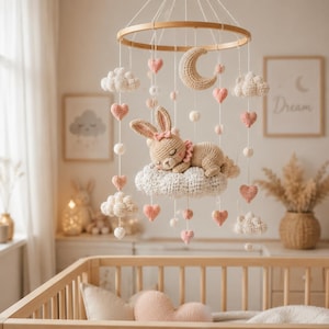 Könnte beinhalten: Ein hängendes Mobile für das Kinderzimmer mit einem runden Holzrahmen, weißen gehäkelten Wolken, rosa gehäkelten Herzen, kleinen weißen Bommeln, einem beigen Halbmond und einem zentralen beigen Häkelhasen, der auf einer großen Wolke schläft.