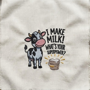 Könnte beinhalten: Ein hellbeiger Stoff mit einer Stickerei, die eine schwarz-weiße Kuh neben einem Milcheimer zeigt. Der Text lautet, I MAKE MILK! WHAT'S YOUR SUPERPOWER? Die Kuh hat eine rosa Schnauze und der Eimer ist mit gelben Akzentlinien versehen.
