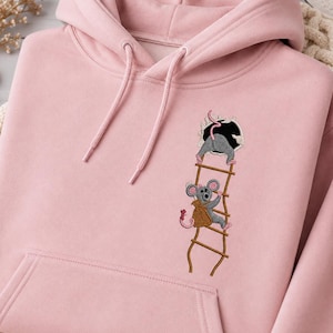 Könnte beinhalten: Ein hellrosa Kapuzenpullover mit einer Stickerei von zwei grauen Mäusen auf einer braunen Leiter. Eine Maus klettert mit einem braunen Sack die Leiter hinauf, während die andere Maus in ein dunkles, kreisförmiges Loch oben an der Leiter kriecht.