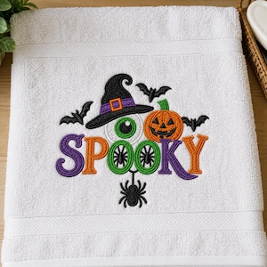Könnte beinhalten: Ein weißes Frotteehandtuch mit einer Stickerei des Wortes SPOOKY in Lila und Orange. Das Motiv zeigt einen schwarzen Hexenhut, einen grünen Augapfel, einen orangefarbenen Kürbis, eine schwarze Spinne und mehrere schwarze Fledermäuse.