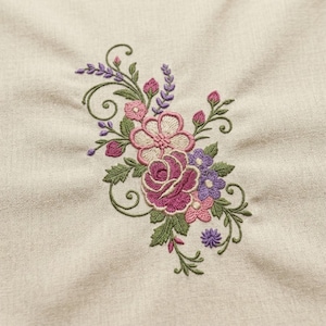 Könnte beinhalten: Ein beigefarbener Stoff mit einer zentralen Maschinenstickerei in Form eines Blumenstraußes. Das Muster besteht aus rosa und violetten Blüten, grünen Blättern und zarten geschwungenen Stielen, die detailliert auf das Textil gestickt sind.