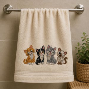 Könnte beinhalten: Ein beiges Handtuch mit einer Stickerei von vier Kätzchen, die in einer Reihe sitzen. Die Kätzchen umfassen einen orangefarbenen Tabby, eine schwarz-weiße Katze, einen grauen Tabby mit herausgestreckter Zunge und ein braun-weißes Siam-Kätzchen.
