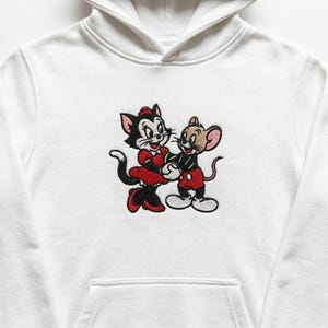 Könnte beinhalten: Ein weißer Kapuzenpullover mit einer zentralen Stickerei, die eine schwarze Katze in einem roten Kleid und eine braune Maus in einer roten Hose zeigt. Die beiden Zeichentrickfiguren halten sich an den Händen, die Katze links und die Maus rechts.