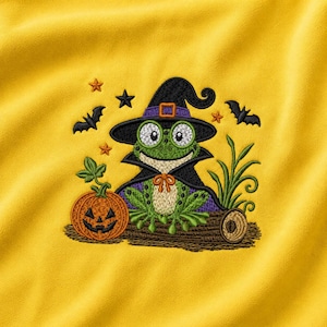 Pode incluir: Um tecido amarelo brilhante com um design bordado de um sapo verde usando um chapéu de bruxa e uma capa preta. O sapo está sentado num tronco castanho ao lado de uma abóbora laranja, rodeado por pequenos morcegos pretos e estrelas.