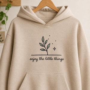 Könnte beinhalten: Ein beigefarbener Kapuzenpullover mit einer Stickerei auf der Brust. Das Motiv zeigt eine kleine grüne Pflanze mit Blättern, einen vierzackigen Stern und drei Punkte über einer horizontalen Linie, darunter der Text enjoy the little things.