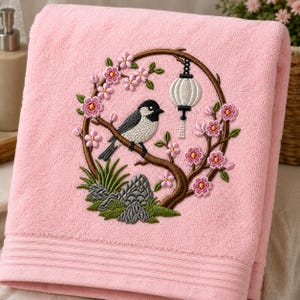 Könnte beinhalten: Ein weiches, rosafarbenes Badetuch mit einer kreisförmigen Stickerei. Das Motiv zeigt einen schwarz-weißen Vogel auf einem braunen Ast, umgeben von rosa Kirschblüten, grünem Gras, grauen Steinen und einer hängenden weißen Papierlaterne.