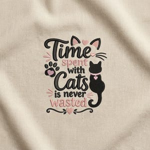 Könnte beinhalten: Ein gesticktes Motiv auf beigem Stoff mit der Silhouette einer schwarzen Katze, die ein rosa Halsband mit Herzanhänger trägt. Der Text Time spent with Cats is never wasted ist in Schwarz und Rosa gehalten, ergänzt durch eine Pfote und kleine Herzen.