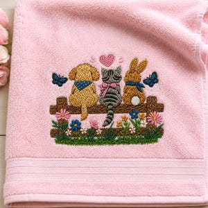 Könnte beinhalten: Ein weiches rosa Handtuch mit einer gestickten Szene, die einen goldenen Hund, eine graue getigerte Katze und einen braunen Hasen auf einem Holzzaun zeigt. Das Design umfasst ein rosa Herz, blaue Schmetterlinge und bunte Blumen am unteren Rand.