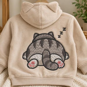 Könnte beinhalten: Ein beigefarbener Kapuzenpullover mit langen Ärmeln aus Plüschmaterial und Katzenohren an der Kapuze. Auf der Rückseite befindet sich eine große Stickerei einer schlafenden grauen getigerten Katze mit eingerolltem Schwanz und ZZZ-Buchstaben.