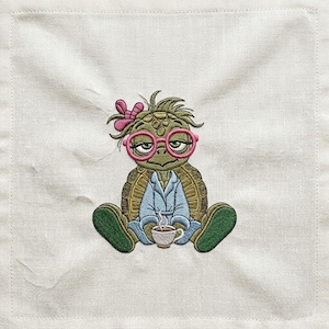 Könnte beinhalten: Ein quadratischer, bestickter Stoffaufnäher mit einer grünen Schildkröte, die eine rosa Brille und einen blauen Morgenmantel trägt sowie eine rosa Schleife auf dem Kopf hat. Die Schildkröte hält eine kleine weiße Kaffeetasse mit Dampf.