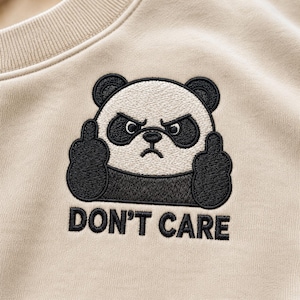 Könnte beinhalten: Ein beigefarbenes Sweatshirt mit einer Stickerei eines wütenden Pandas mit schwarz-weißem Fell. Der Panda hebt beide Hände und zeigt den Mittelfinger. Unter dem Panda ist der Text DON'T CARE in schwarzer Schrift aufgestickt.