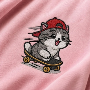 Könnte beinhalten: Ein hellrosa Stoff mit einer Stickerei, die eine grau-weiße getigerte Katze mit roter Baseballkappe auf einem schwarzen Skateboard mit gelben Rollen zeigt. Die Katze hat einen fröhlichen Gesichtsausdruck mit offenem Mund und sichtbarer Zunge.