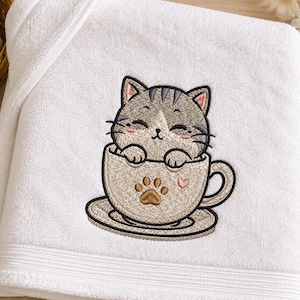 Könnte beinhalten: Ein weißes Baumwollhandtuch mit einer Stickerei, die eine graue getigerte Katze in einer beigen Teetasse zeigt. Die Katze hat geschlossene Augen, rosa Wangen und Schnurrhaare. Die Tasse ist mit einer braunen Pfotenabdruck und einem kleinen rosa Herz verziert.