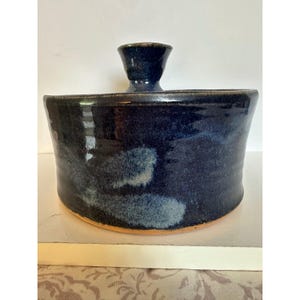 May include: A cylindrical ceramic container with a deep navy blue glaze featuring lighter blue mottled accents. The vessel has a smooth, glossy finish and a central, flared handle on top, with a visible unglazed terracotta-colored base rim.