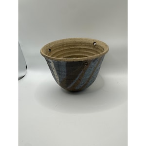 May include: A speckled ceramic hanging planter with a wide, flared rim and three small holes for suspension. The exterior features a textured, multi-colored glaze in shades of dark brown, slate blue, and cream, while the interior is light beige.