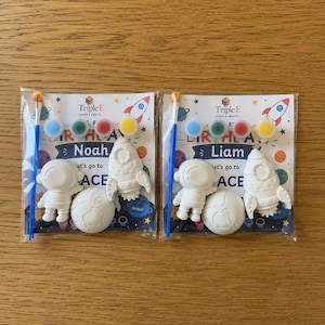 May include: Two space-themed craft kits in clear plastic packaging, each containing a white plaster astronaut, rocket, and planet, a blue paintbrush, and four small paint pots. The cards feature the text Triple E Paints & Crafts, Birthday, and Let's go to SPACE, with personalized names Noah and Liam.