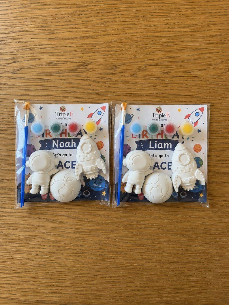 May include: Two space-themed craft kits in clear plastic packaging, each containing a white plaster astronaut, rocket, and planet, a blue paintbrush, and four small paint pots. The cards feature the text Triple E Paints & Crafts, Birthday, and Let's go to SPACE, with personalized names Noah and Liam.