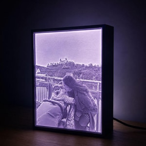 Könnte beinhalten: Ein rechteckiger Leuchtkasten mit einem hinterleuchteten, einfarbig violetten Foto einer Person, die sich zu einem Kind im Kinderwagen beugt, mit einer Burg auf einem Hügel im Hintergrund. Der Rahmen ist schwarz und ein Kabel ragt seitlich heraus.