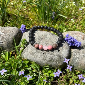May include: A black lava stone bead bracelet with pink and grey gemstone accents. The bracelet is displayed on a grey stone surrounded by green grass and small purple flowers. A natural, earthy accessory.