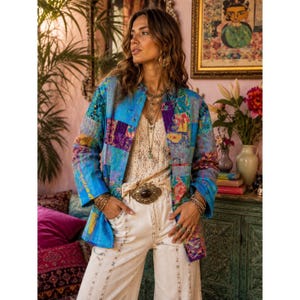 Könnte beinhalten: Eine Patchwork-Jacke in Blau-, Lila- und Gelbtönen. Die Jacke hat ein kragenloses Design und lange Ärmel. Das Model trägt ein cremefarbenes Spitzentop und eine weiße Hose. Die Jacke ist im Bohemian-Stil gehalten.