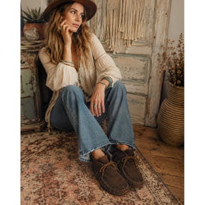 May include: A cream-colored long-sleeve blouse with delicate embroidery, paired with blue denim flare jeans and dark brown suede ankle boots featuring fringe details and lace-up closures. The outfit is accessorized with a wide-brimmed brown hat.