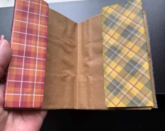 May include: Open notebook with brown paper pages and plaid covers in shades of orange, red, and yellow. The notebook is open, revealing the interior pages. The plaid pattern is a classic design.