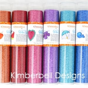 Puede incluir: Una colección de tubos de purpurina Kimberbell Designs en varios colores, incluyendo plata, oro y cobre. Cada tubo está lleno de purpurina y tiene una etiqueta con un diseño. El logotipo de Kimberbell Designs es visible.