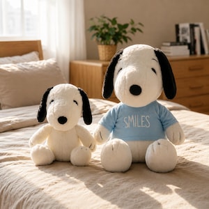 May include: Two plush Snoopy dolls. One is smaller and the other is larger. Both have white fur, black ears, and black eyes. The larger doll wears a light blue shirt with the word "SMILES" printed on it.