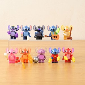 May include: A collection of colorful Stitch character figurines, each in a unique pose and holding different accessories. The figurines are arranged on a clear display shelf, with a light wooden surface in the background.